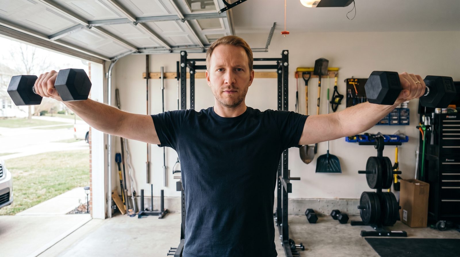 A man in a navy t-shirt doing dumbbell lateral raises in a garage gym with the door open