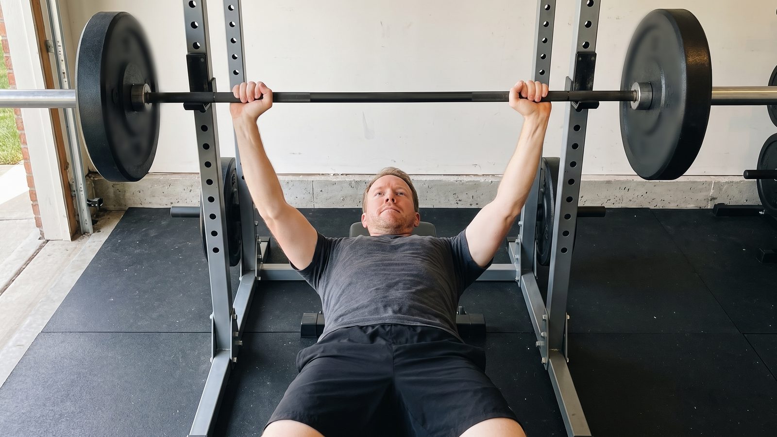 A man at the top of a barbell bench press inside a garage power rack, arms locked out and the bar held over his shoulders, seen from the foot of the bench