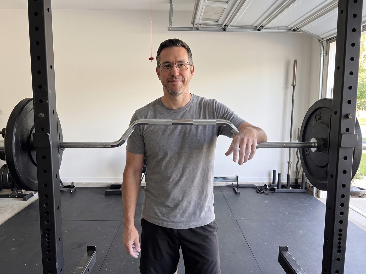 A man in glasses standing beside a power rack in a garage gym, one chalked hand resting on the racked chrome cambered bar, its raised middle section a hand's width above the grips, two plates a side