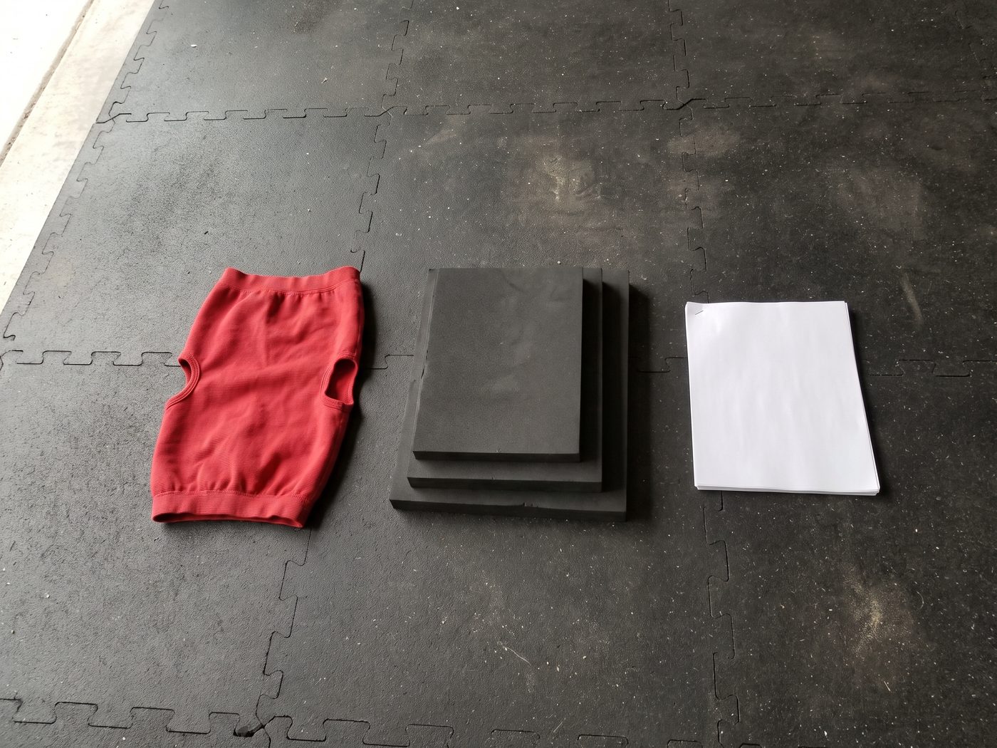 A red elastic bench press sleeve, a stack of three black foam bench boards and a stapled printed program lying face down on a black rubber gym floor