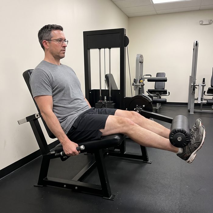 A man in glasses on a leg extension machine, one leg pushing the roller a third of the way up from the hanging position
