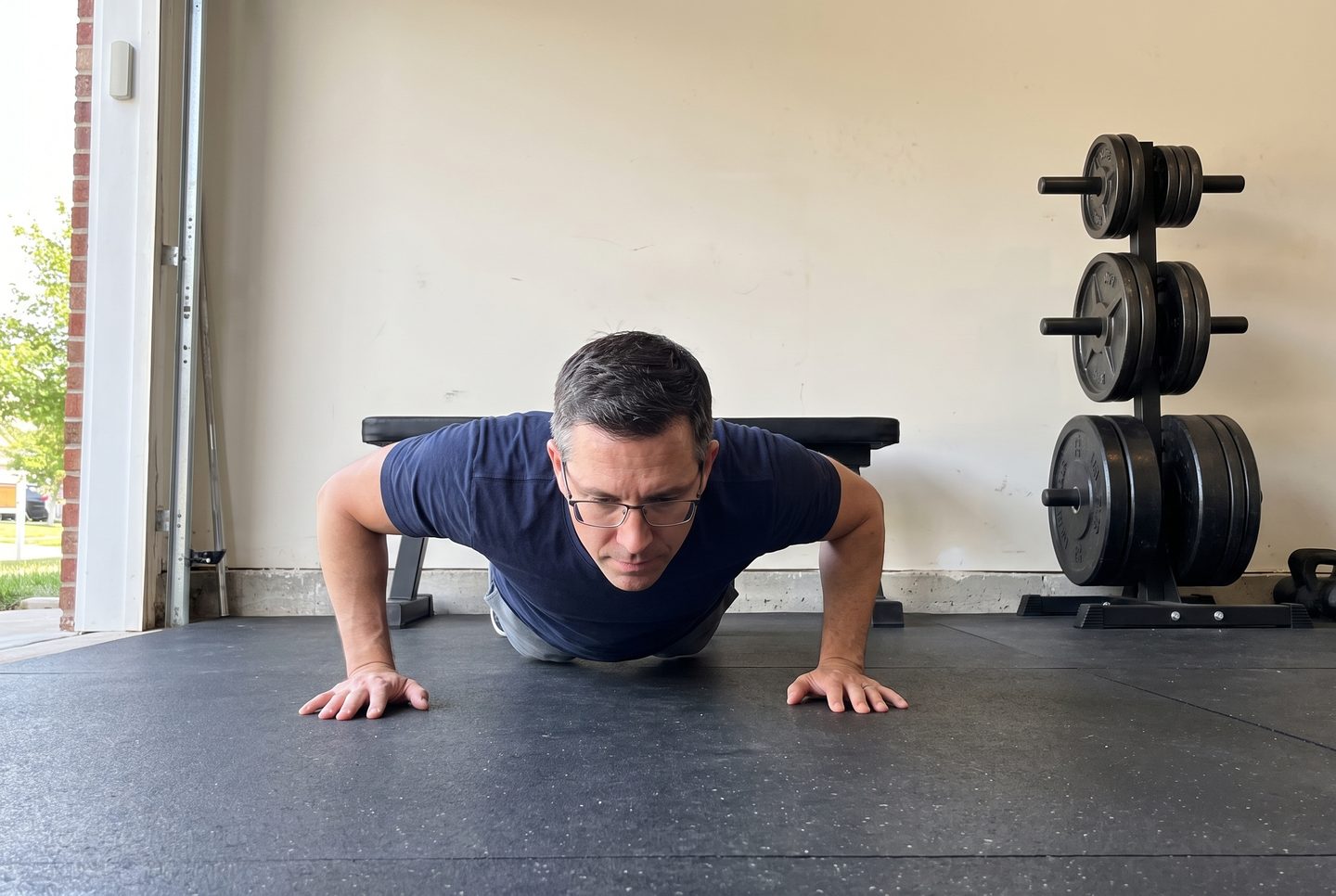 A man in his forties at the bottom of a push-up on a rubber garage floor with a plate tree behind him