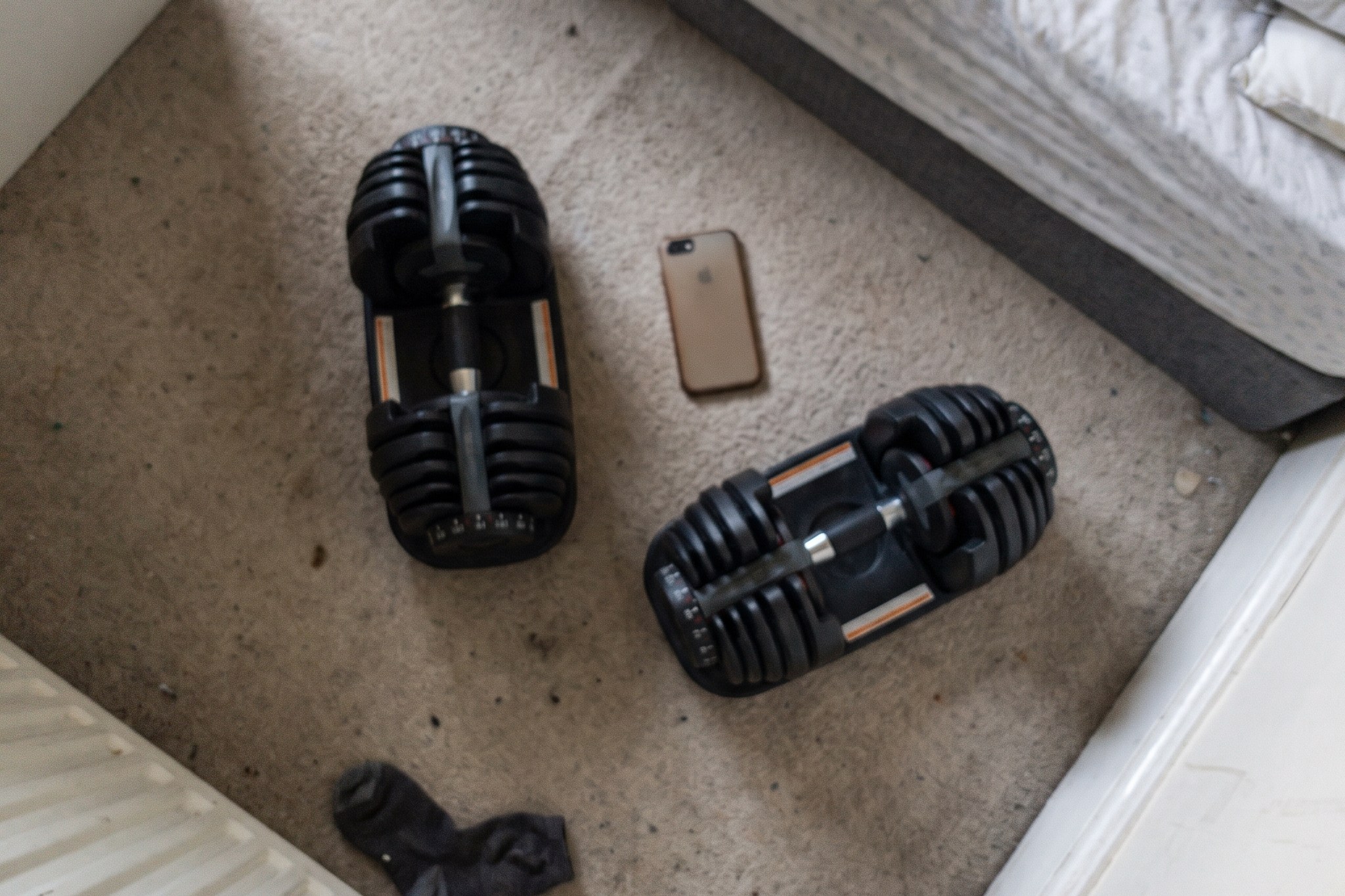 A pair of adjustable dumbbells and a phone on a bedroom carpet beside an unmade bed