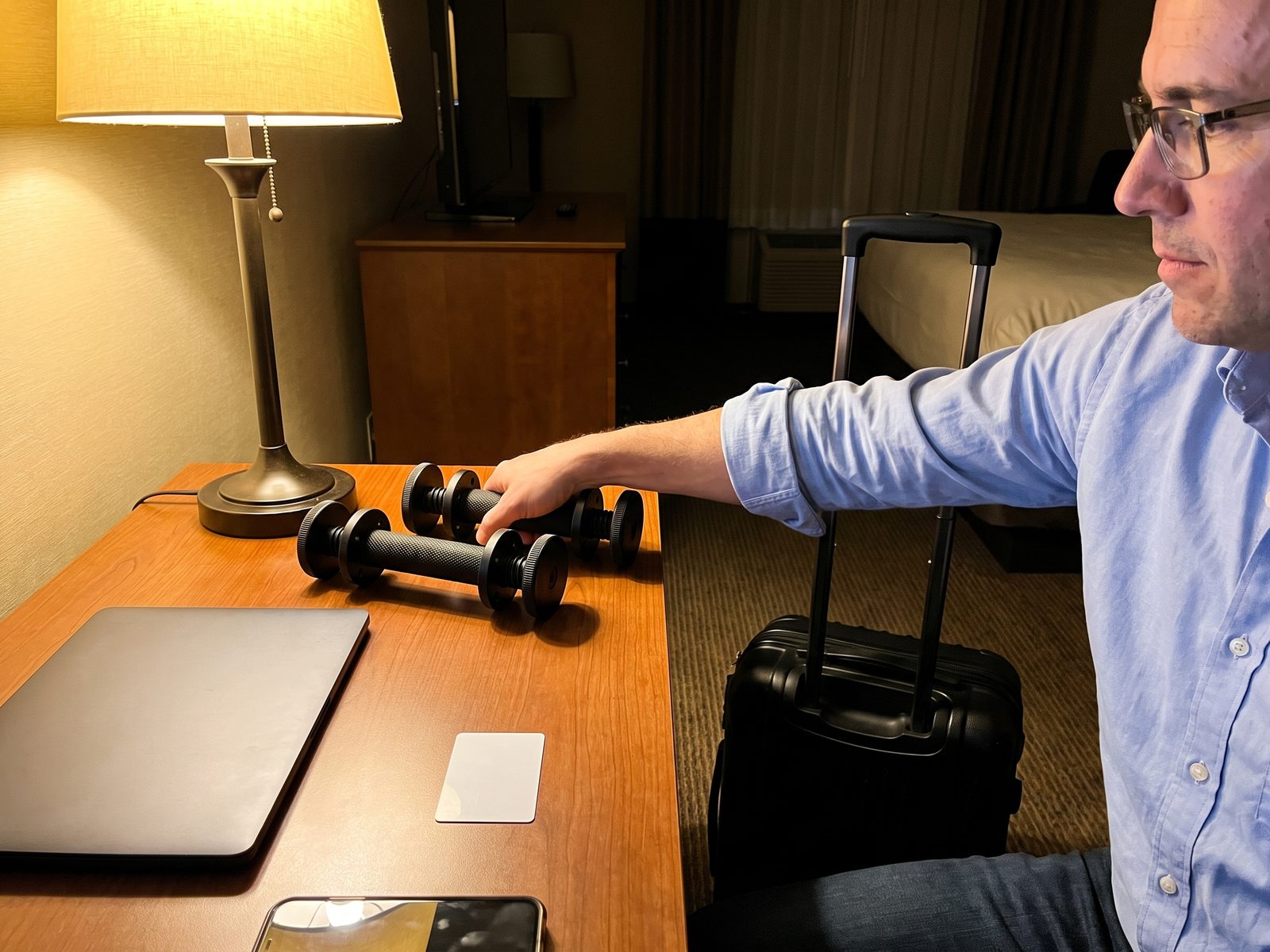 Two black plate loaded dumbbell handles on a hotel room desk beside a closed laptop and a key card, a man's hand in a rolled shirt sleeve reaching for one