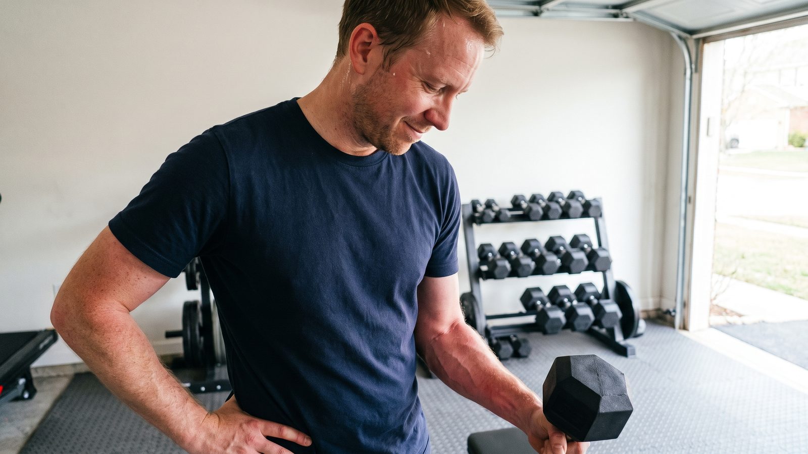 A man in a navy t-shirt standing in a garage gym after a set, one hand on his hip and a small black hex dumbbell in the other, looking down at his flushed, pumped forearm with a tired half-smile, a dumbbell rack behind him