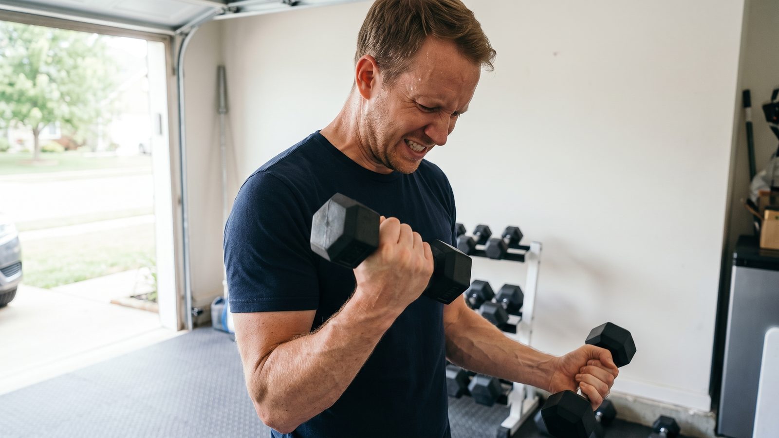 A man in a navy t-shirt standing in a bright garage gym, grinding out the last rep of a long set of curls with a small black hex dumbbell, forearm and biceps pumped, face strained, a dumbbell rack out of focus behind him