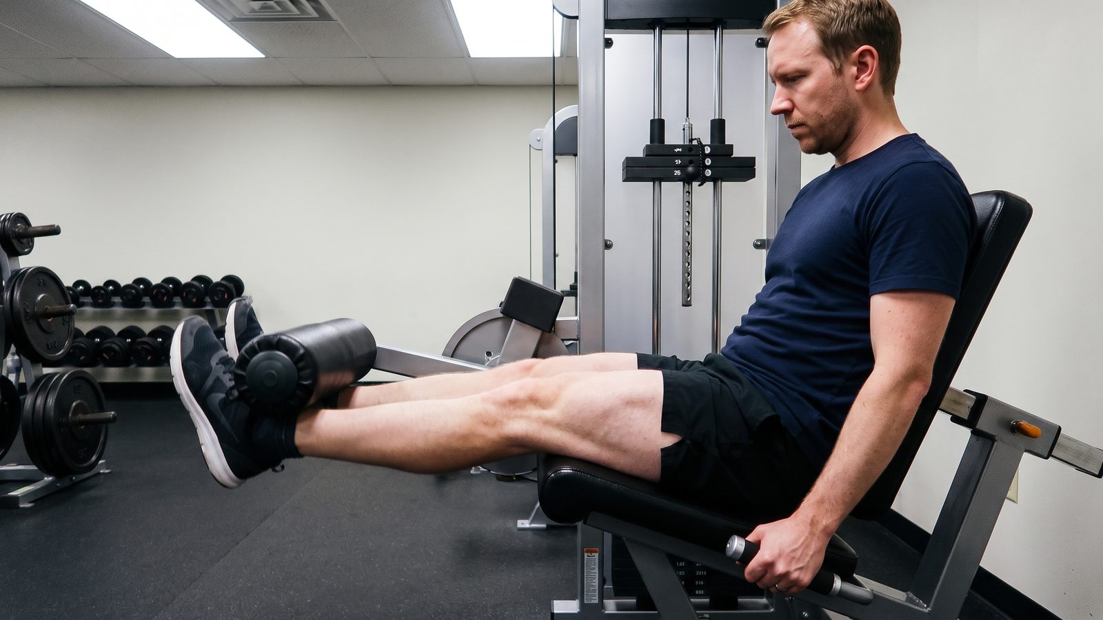 Side view of a man in a navy t-shirt seated on a leg extension machine in a gym, legs almost straight against the roller pad, thighs tight, the pin set high on the weight stack