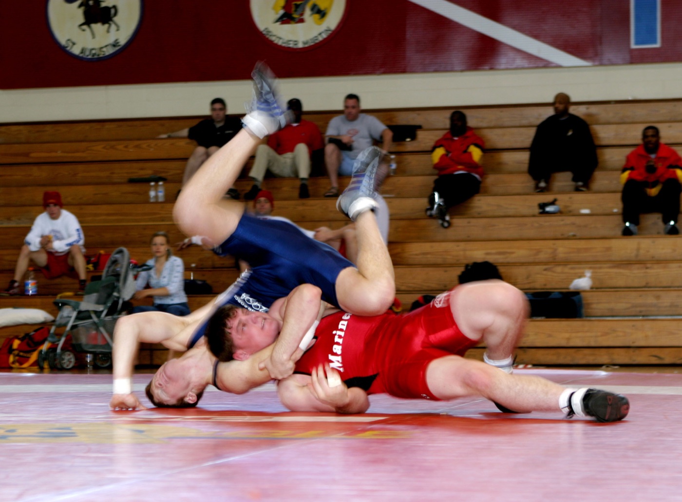A wrestler in a red singlet bridging on his neck with an opponent's full weight driving him into the mat