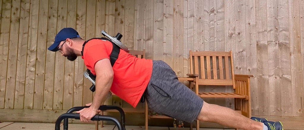 An owner doing a loaded push-up on low handles in a backyard, with plates on the front and back of the vest