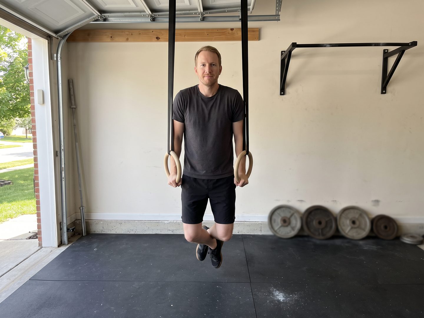 The writer holding the top of a bodyweight ring dip in his garage, with his old iron plates leaning unused against the wall