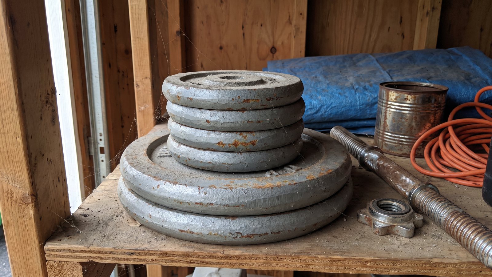 A dusty stack of old grey cast iron weight plates on a garage shelf next to a rusted chrome dumbbell bar and its loose spin collar