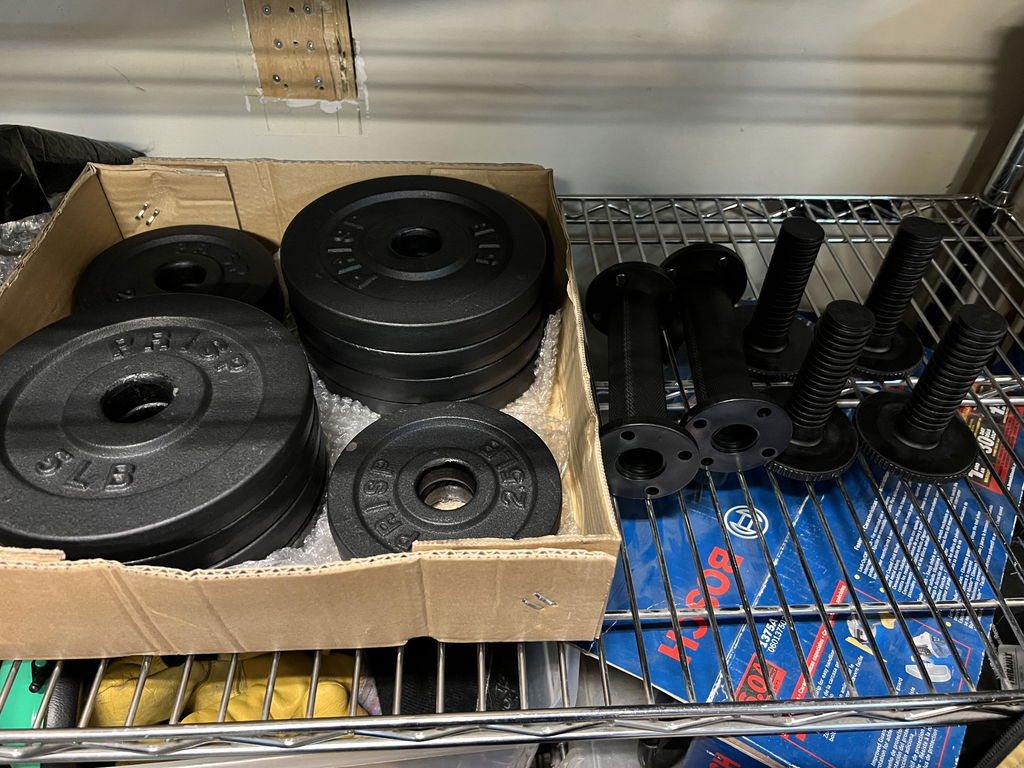 Black plate loaded dumbbell grips and their threaded posts on a wire shelf next to a box of standard plates