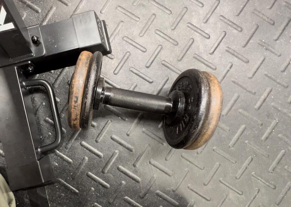 A plate loaded dumbbell handle carrying two worn iron plates on a black rubber gym floor next to a rack