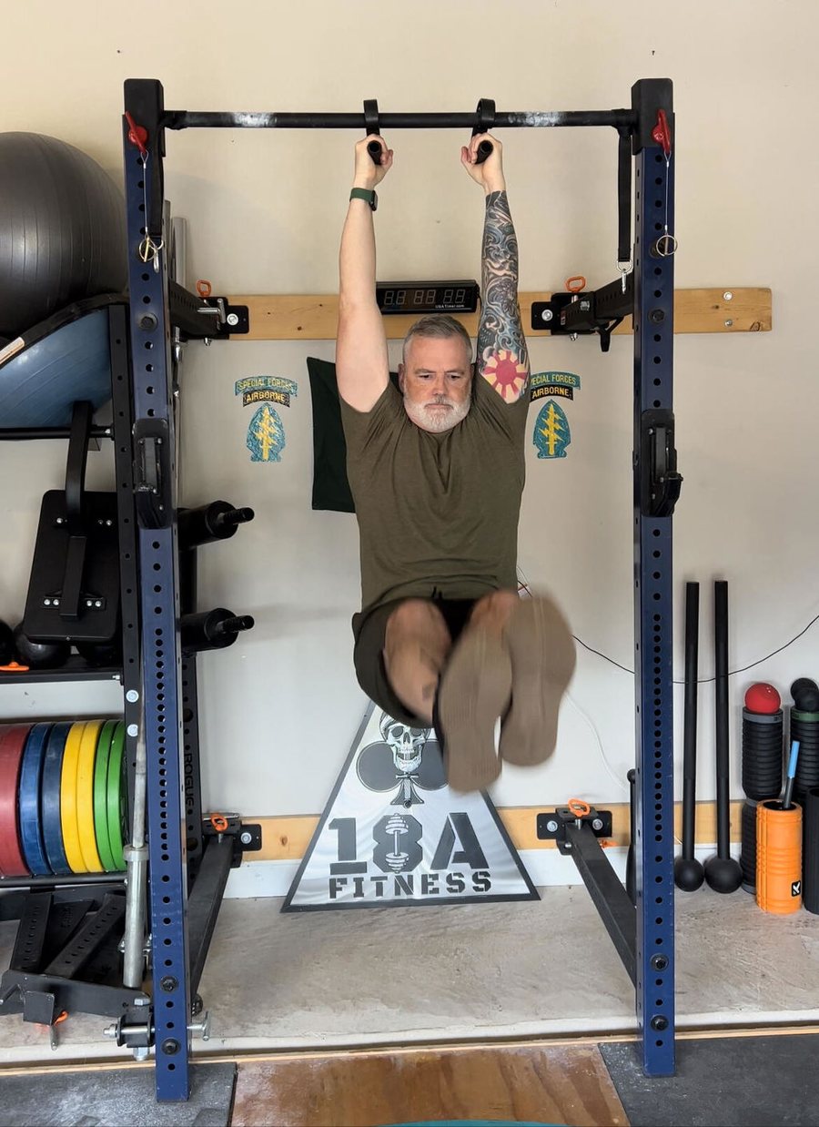 A grey-bearded man hanging from a home rack pull-up bar on a pair of Swissies handles