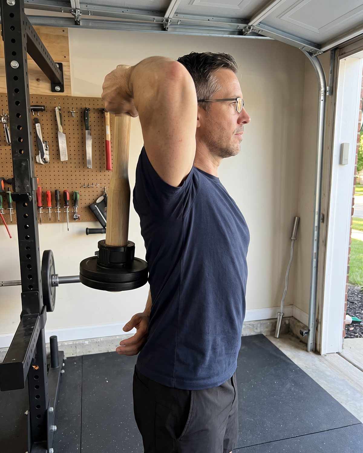 The author in his garage holding the wooden club over his head so it hangs straight down behind his back, two small black plates on the end