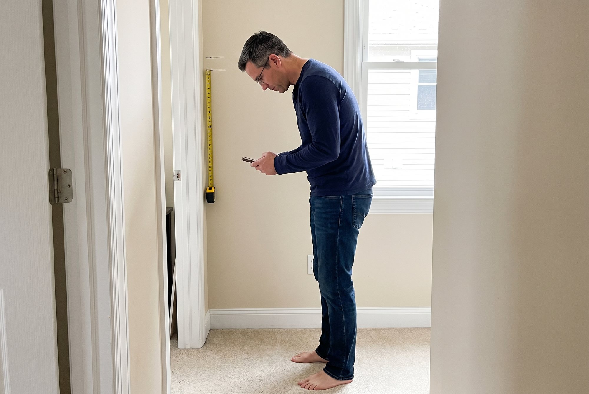 Side view of a man in a navy shirt reading his phone in a hallway, head forward and shoulders rounded, a yellow tape measure and two pencil marks on the white door frame beside his head