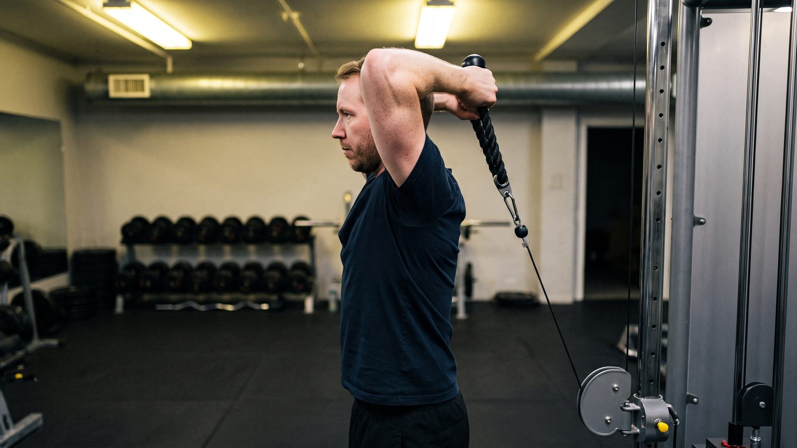 Side view of a man in a navy t-shirt in a gym doing an overhead cable triceps extension, both upper arms pointing up beside his head, a rope attachment held behind his head and the cable running down behind his back to a low pulley